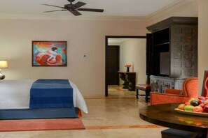 A hotel room with a bed, a television, a dining table with fruit, and a painting on the wall.