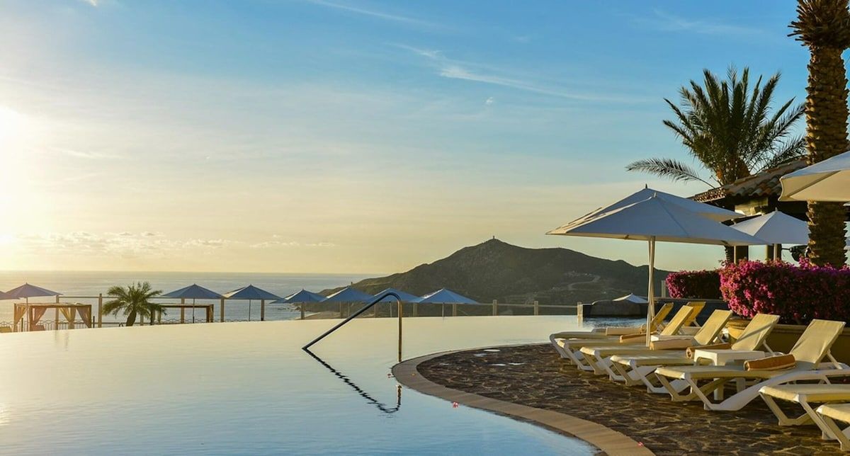 Zona de piscina con sillones de descanso y sombrillas, con vistas a un paisaje montañoso.