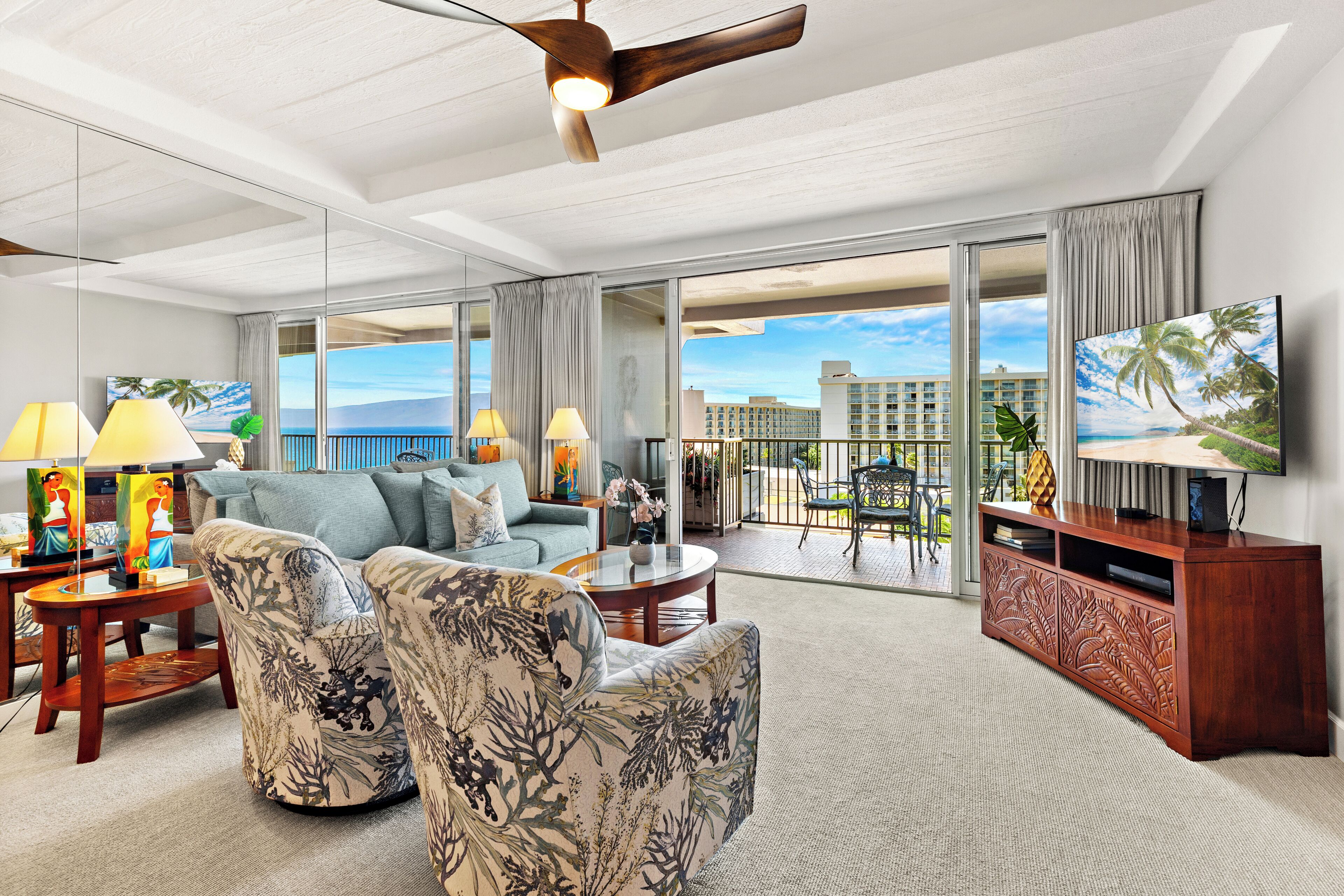 A spacious living room with a large sofa, two armchairs, a coffee table, and a TV on a wooden cabinet. The room has large windows offering a view of the ocean and a building.