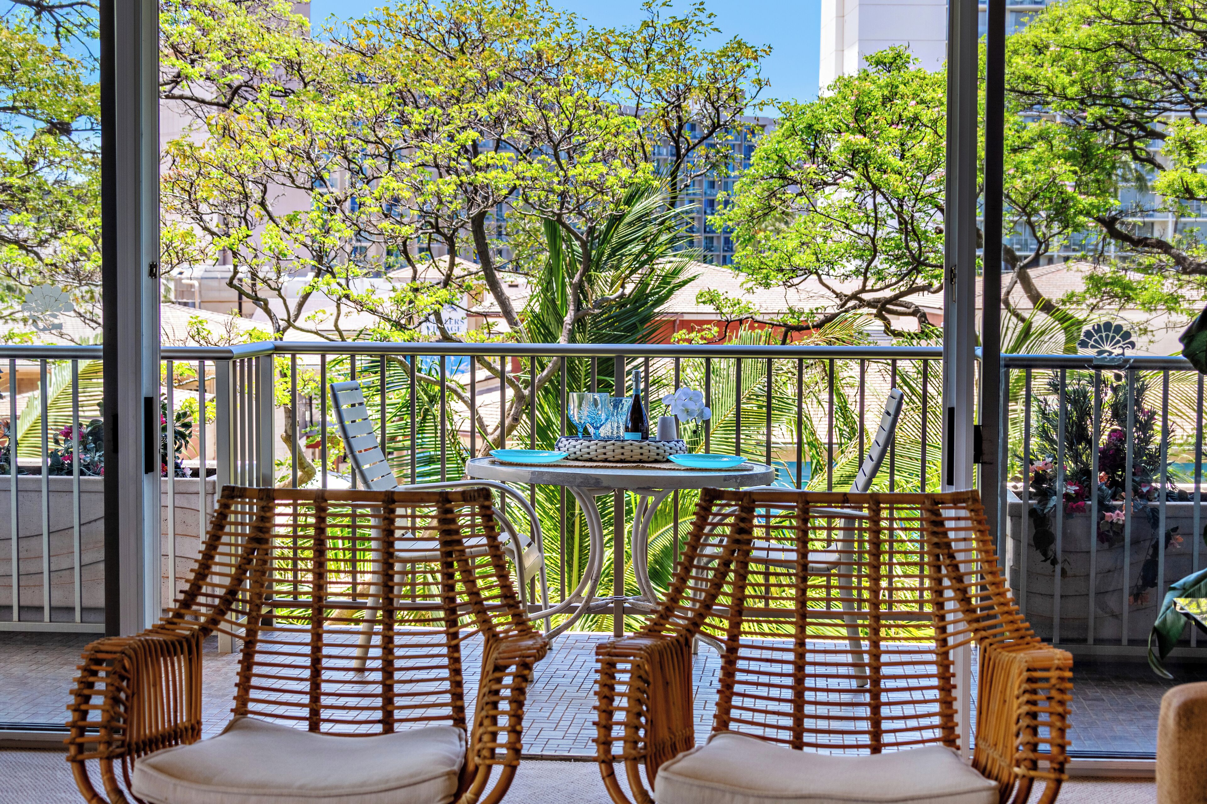 A balcony with wicker chairs and a table, overlooking a garden with trees and a building in the background.