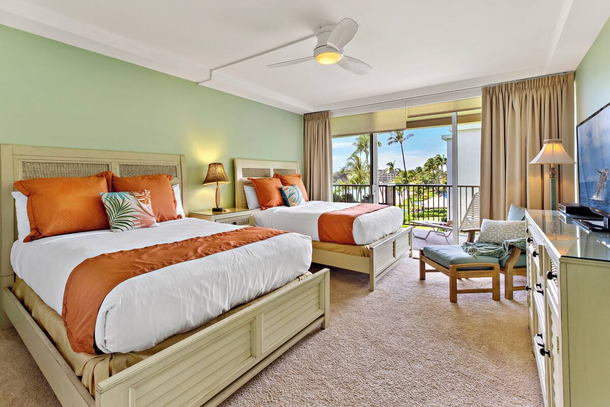 Une chambre d’hôtel avec deux lits, un balcon offrant une vue, un bureau avec une télévision et un ventilateur de plafond.