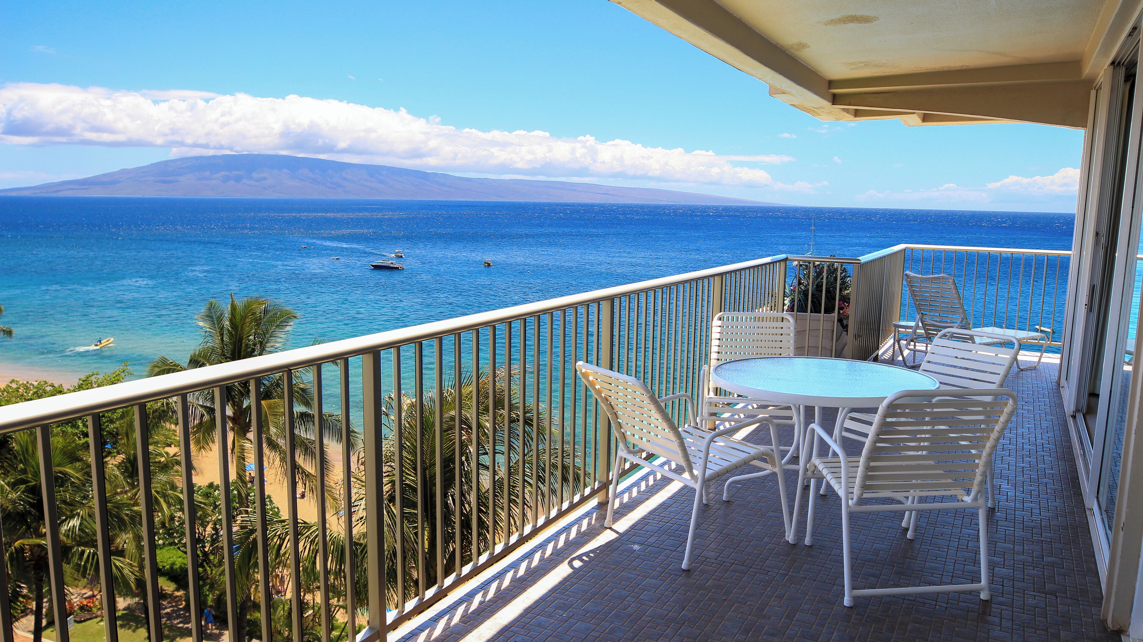 A balcony with a table and chairs, overlooking a beach and ocean with boats.