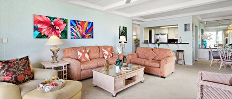 A spacious living room with a sofa, ottoman, and coffee table, a kitchen in the background, and a ceiling fan.