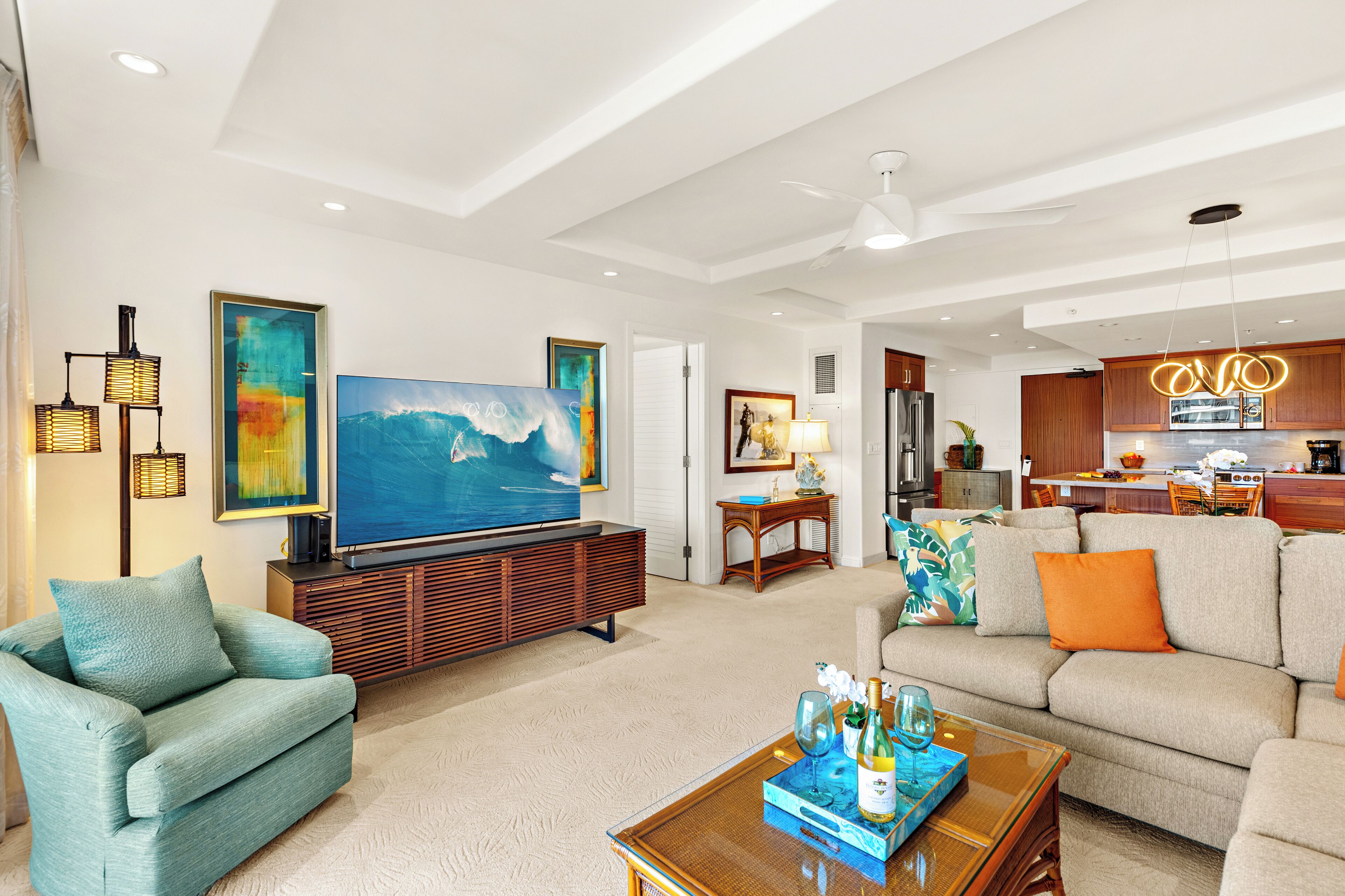 A spacious living room with a large flat-screen TV, comfortable seating, and a kitchen area visible through an open door.