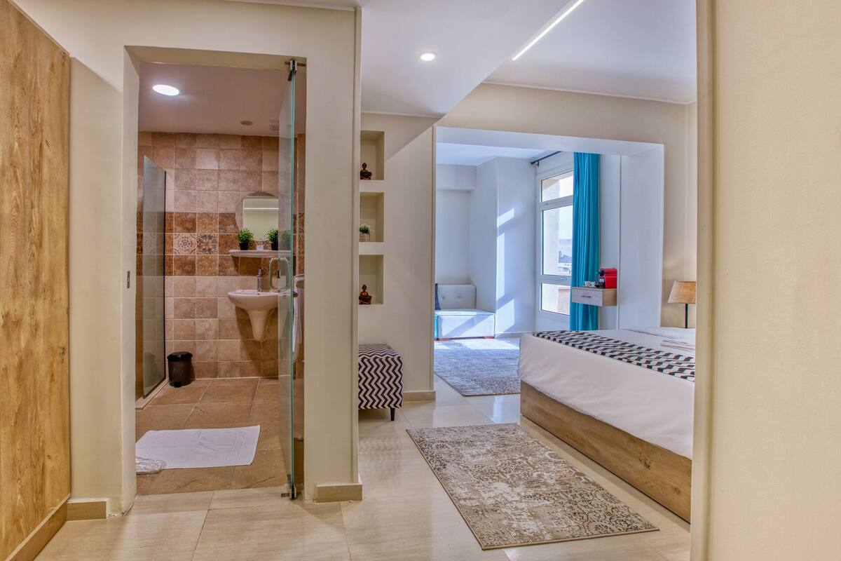 A modern bathroom with a glass shower enclosure, a sink, and a vanity. There is a bed with a patterned bedspread and a bedside table with a lamp. The room has a wooden floor and a view of the city through a window with blue curtains.