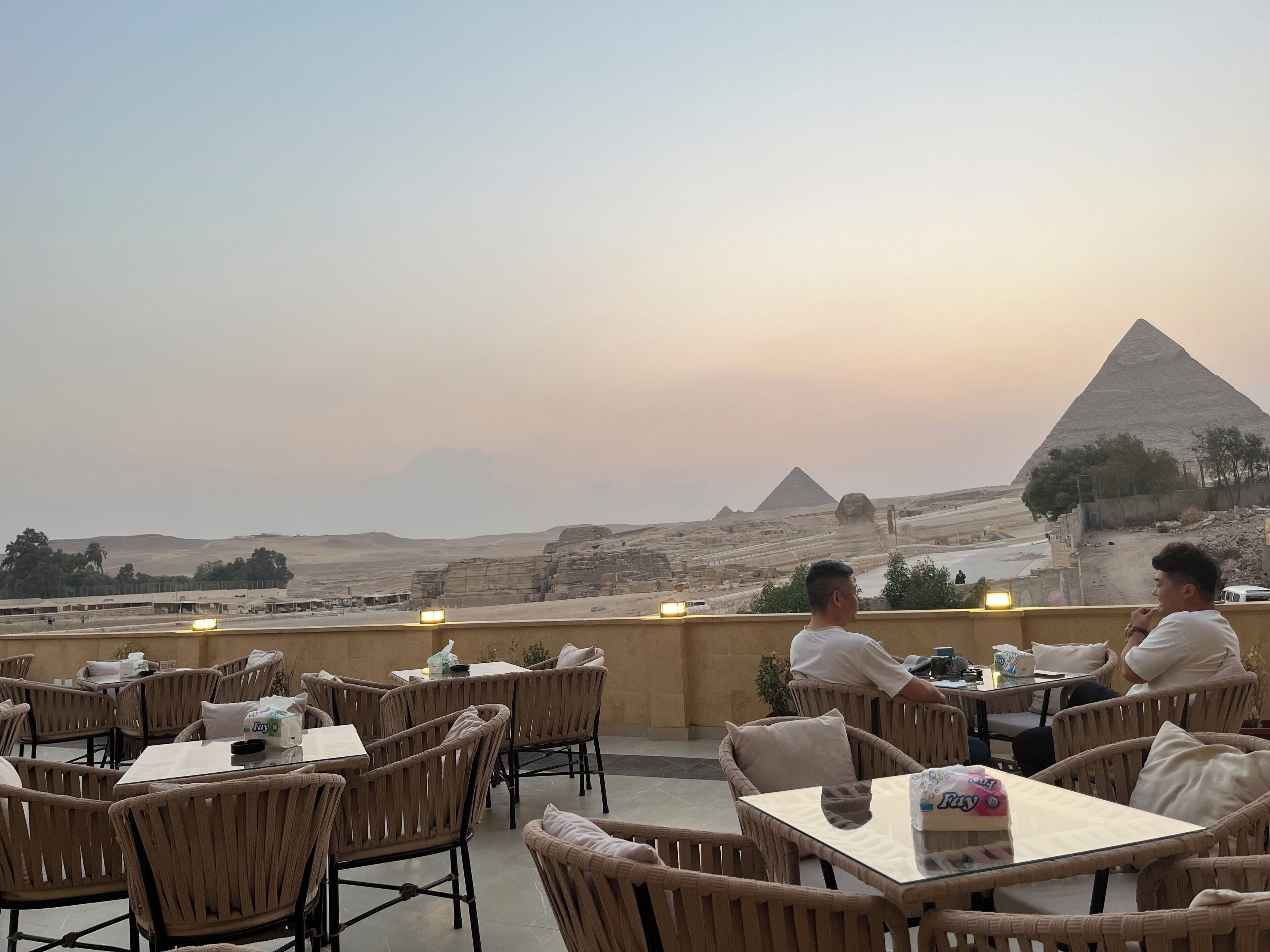 A terrace with wicker furniture and tables, overlooking a desert landscape with pyramids and a sphinx.