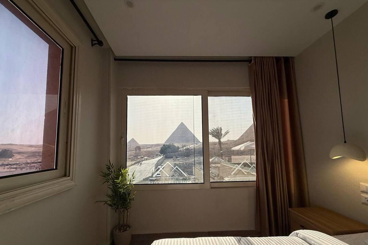 A hotel room with a large window overlooking the pyramids, a bed, a bedside table, a lamp, and a potted plant.