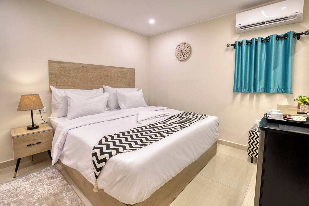 A neatly arranged hotel room with a large bed, a bedside table with a lamp, a small table with a tray of breakfast items, and a wall-mounted air conditioner.