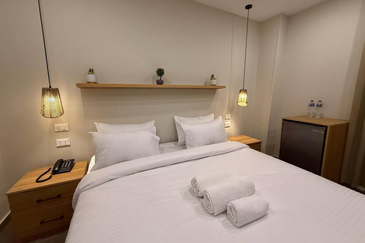 A modern hotel room with a neatly made bed, bedside lamps, a small shelf with plants, a desk with a phone, and a minibar.