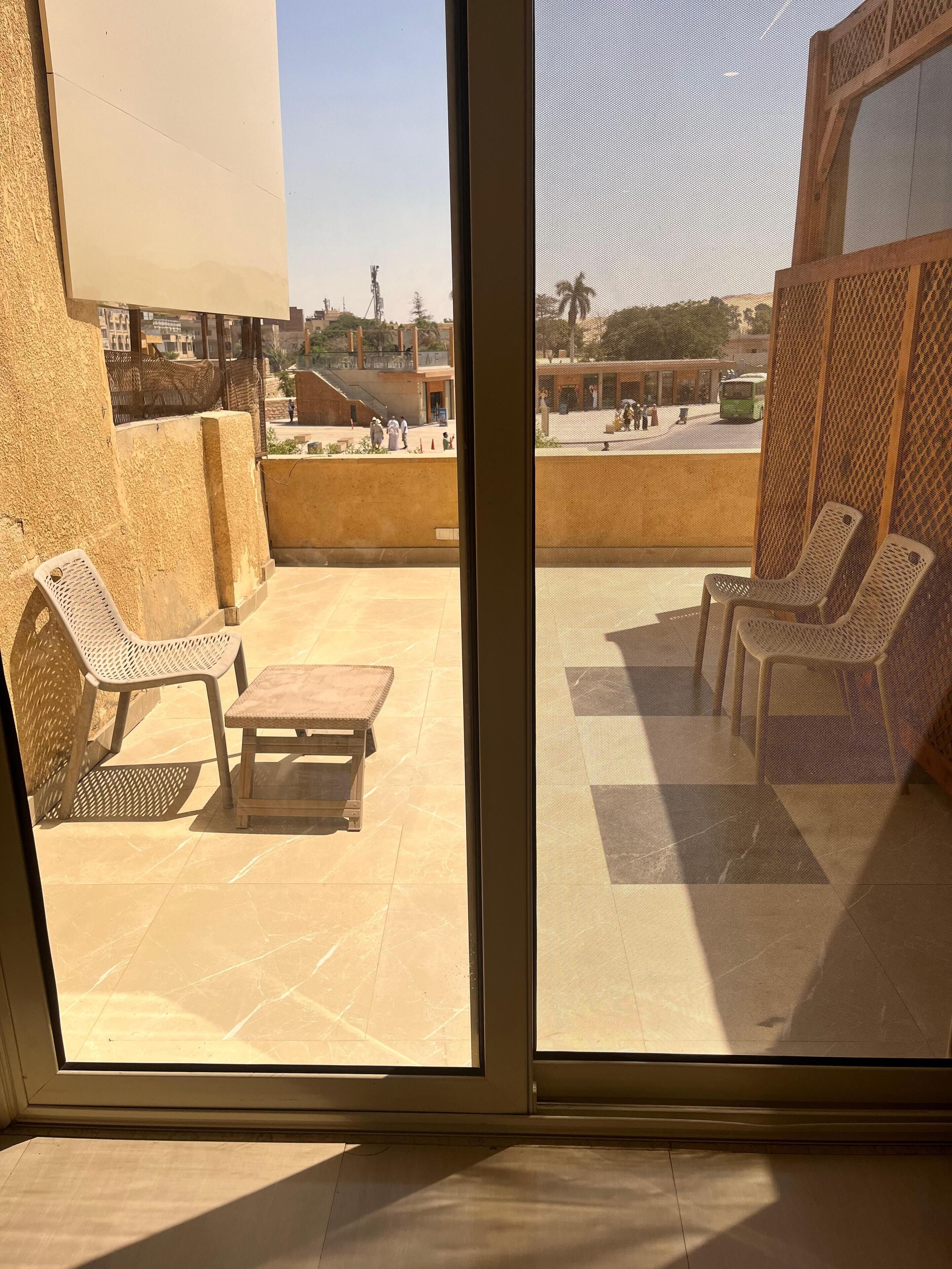 A balcony with a table and chairs, a view of a street with people and a bus, and a building in the background.