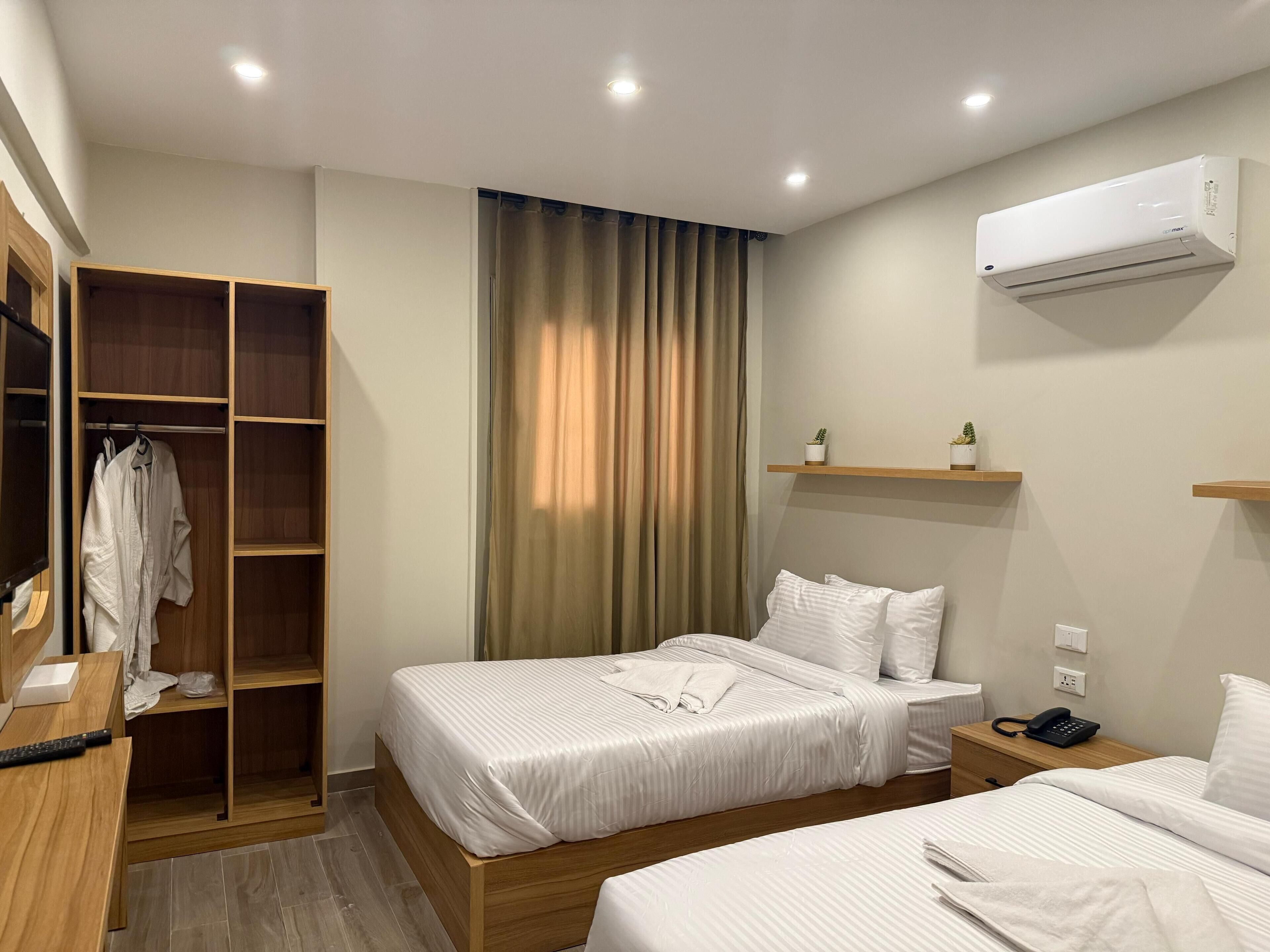 A hotel room with two beds, a wooden wardrobe, a bedside table with a phone, and a wall-mounted air conditioner.