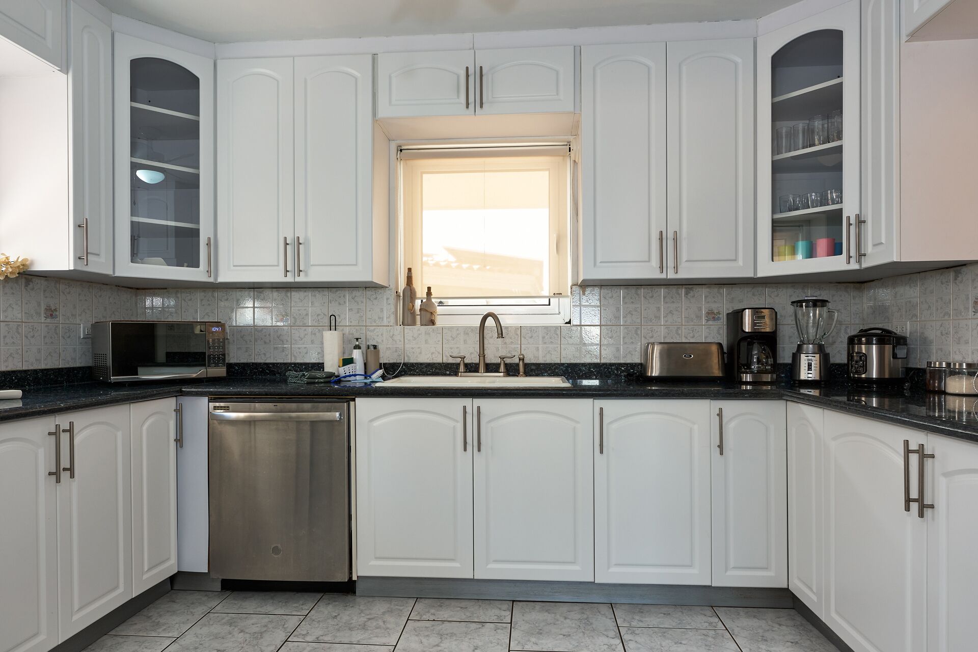 A modern kitchen with white cabinetry, a stainless steel dishwasher, a built-in oven, and a window above the sink.
