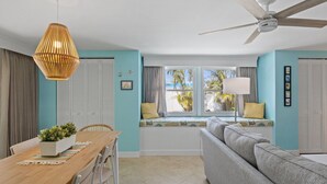 A room with a dining table, chairs, a sofa, a ceiling fan, and a window with a view of palm trees. - Da Beach House: 2 Blocks to Beach, Pool & Spa by Anna Maria Life Vacation Rentals (Holmes Beach)