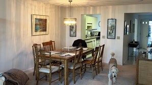 Un coin repas avec une table et des chaises en bois, un lustre et un chien qui passe.