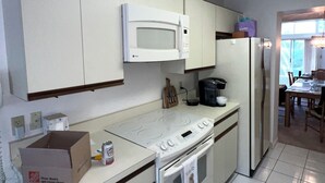 Une cuisine avec des meubles blancs, un four à micro-ondes, une cuisinière et un réfrigérateur. Il y a une boîte sur le comptoir et une canette de soda.