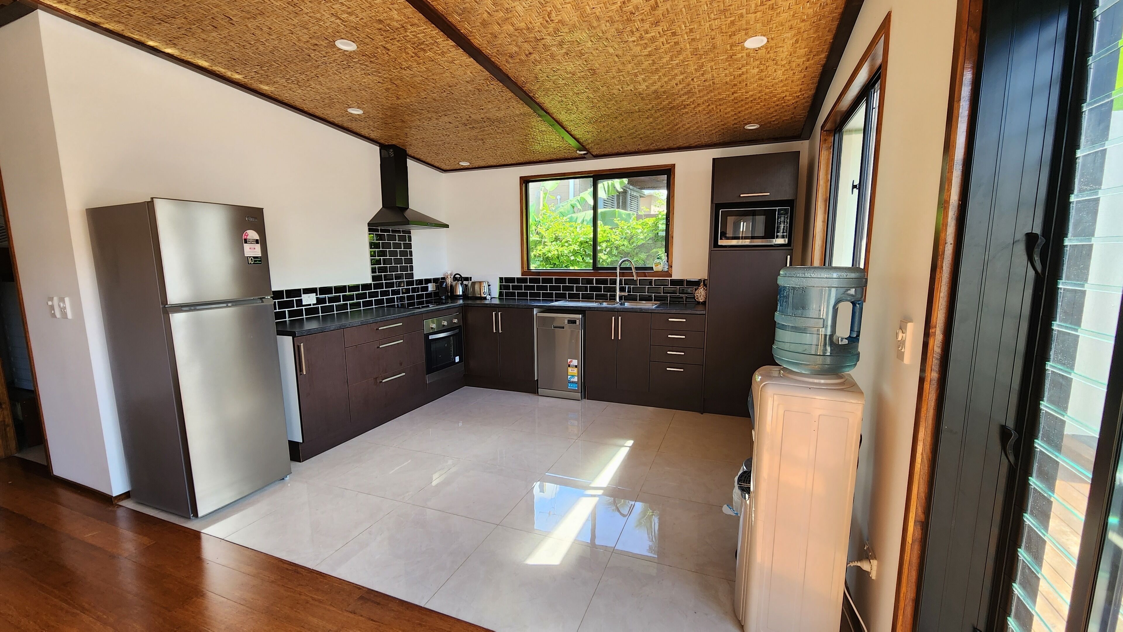 A modern kitchen with dark cabinetry, a stainless steel refrigerator, a dishwasher, and a large window providing a view of greenery outside.