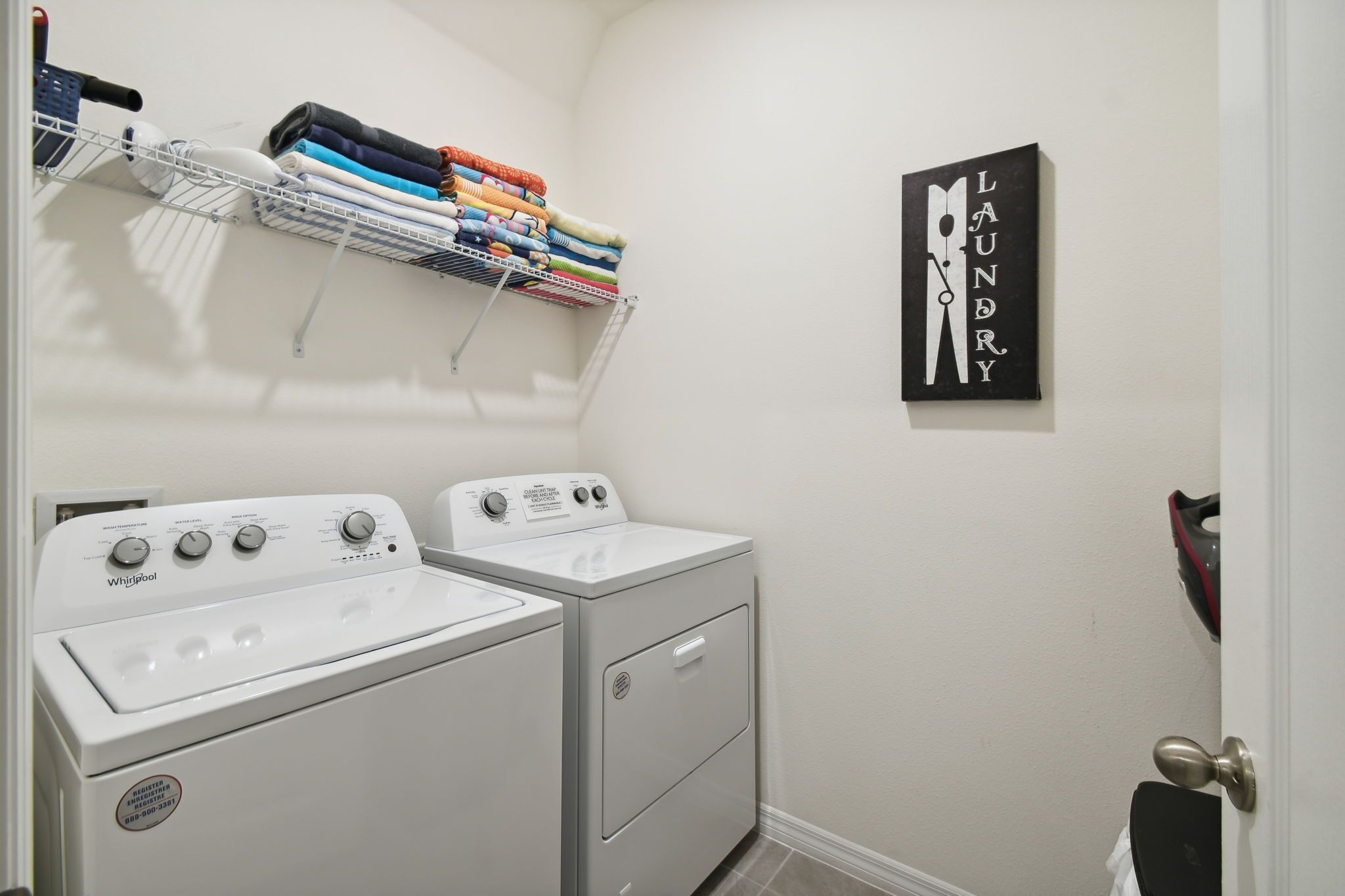 A laundry room with two Whirlpool washers and dryers, a drying rack with folded clothes, and a wall-mounted sign that reads "Laundry".