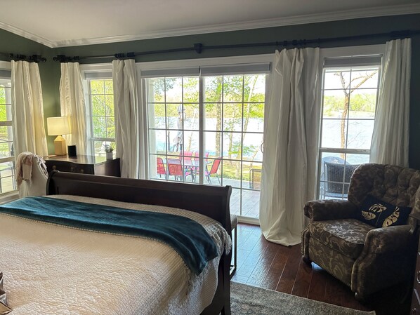 A bedroom with a bed, a chair, a desk with a lamp, and large windows with curtains.