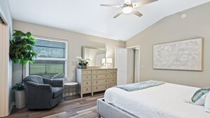 Quarto com cama, cômoda, cadeira, ventilador de teto, janela com persianas e uma planta.