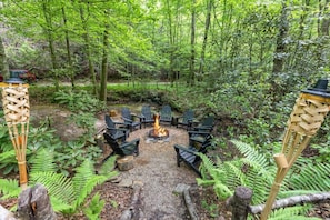A forested area with a fire pit surrounded by chairs.