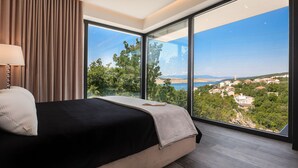 A modern bedroom with a large bed, a nightstand, and a view of a coastal landscape through floor-to-ceiling windows. - Luxury Villa Cloer, Jadranovo/Crikvenica (Jadranovo)