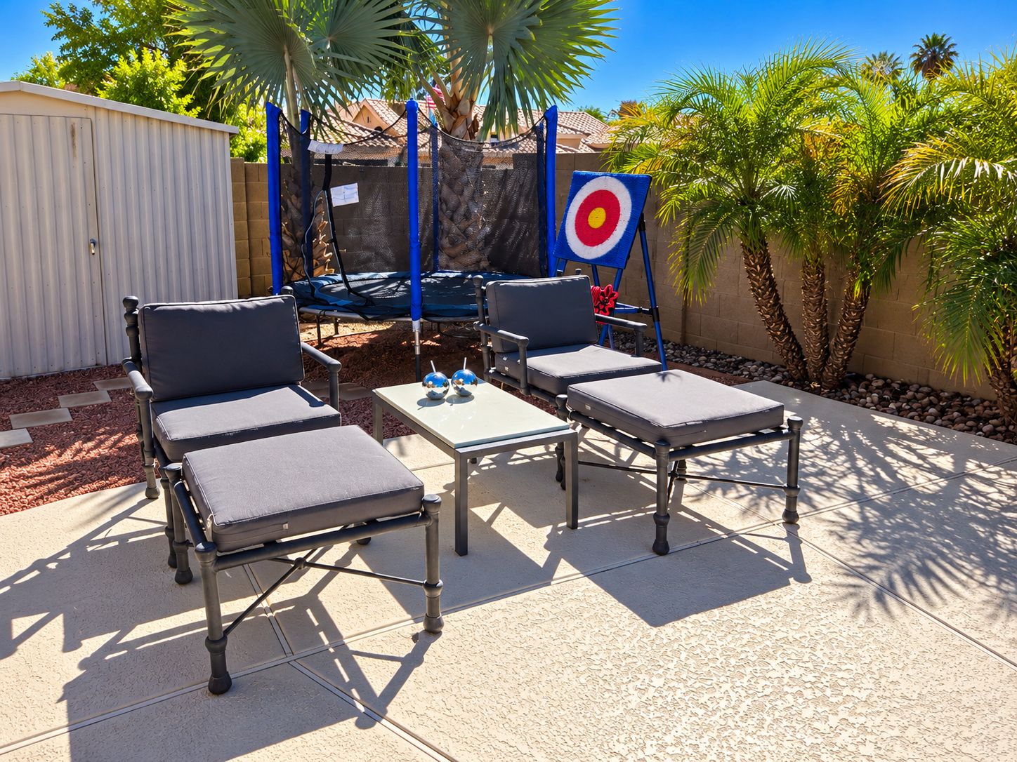 An outdoor seating area with two lounge chairs and a small table, surrounded by palm trees and a fenced area.