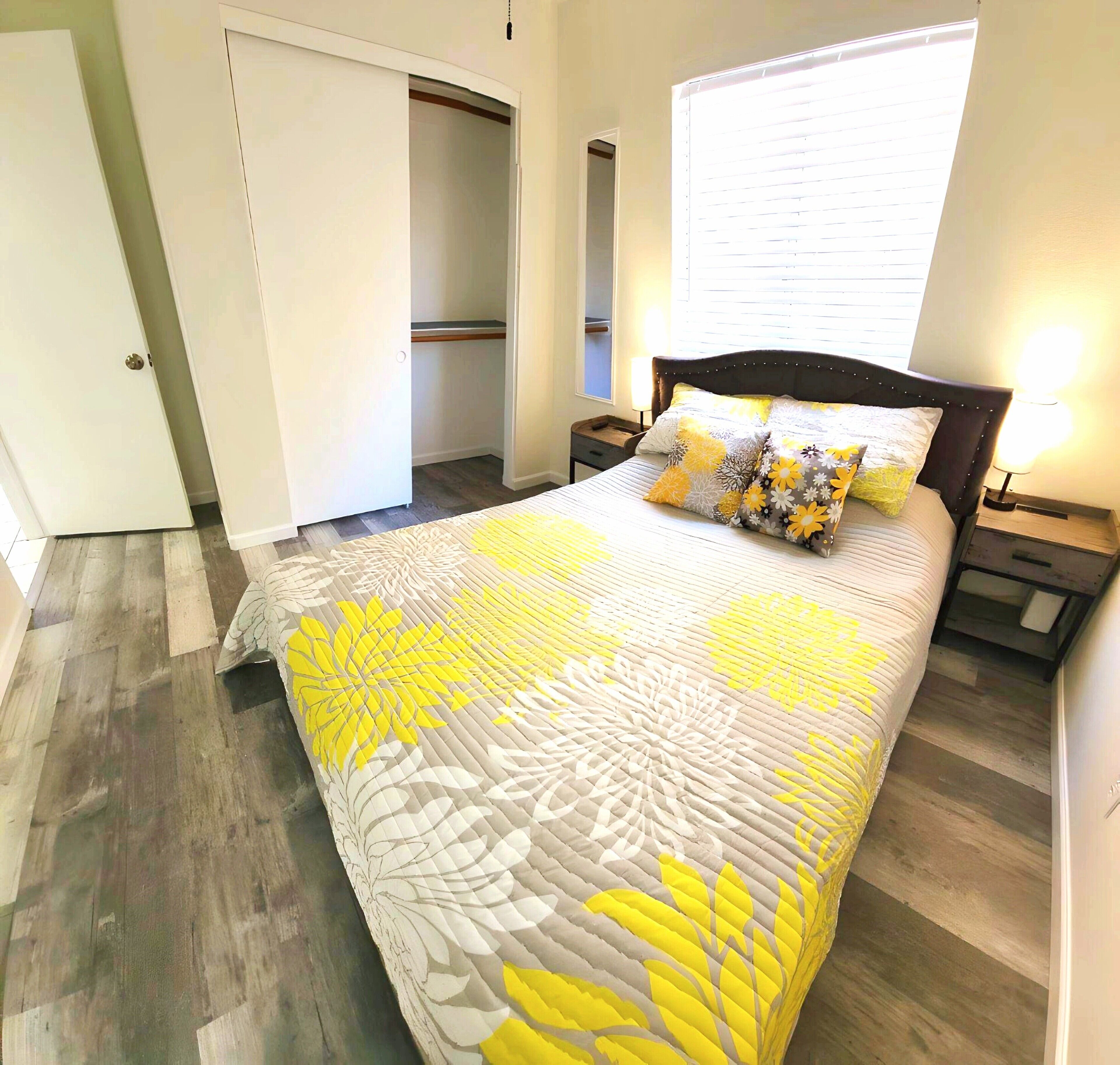 A bedroom with a bed, a nightstand, a bedside table, a lamp, and a window with blinds.