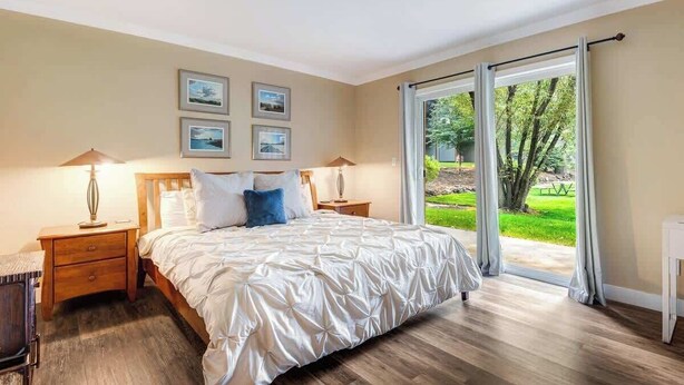 Un dormitorio con una cama grande, dos mesitas de noche con lámparas, vistas a un jardín a través de un ventanal corredero y cuadros enmarcados en la pared.
