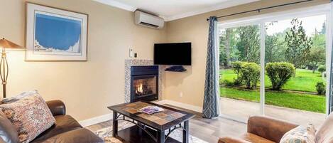 A living room with a fireplace, leather sofas, a coffee table, a flat-screen TV, and a large sliding glass door leading to a garden.