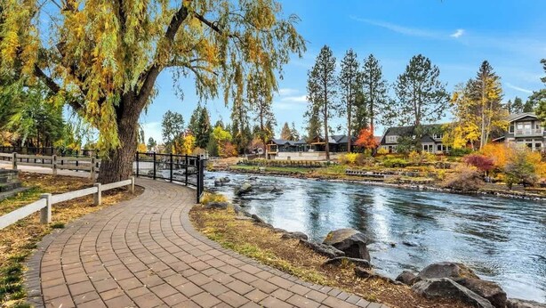 Une promenade dallée longe une rivière, un pont enjambe celle-ci, et l’ensemble est entouré d’arbres aux couleurs automnales et d’immeubles d’habitation.