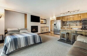 Quarto de hotel moderno com uma cama grande, lareira, área de cozinha com armários de madeira e um banco de bar.