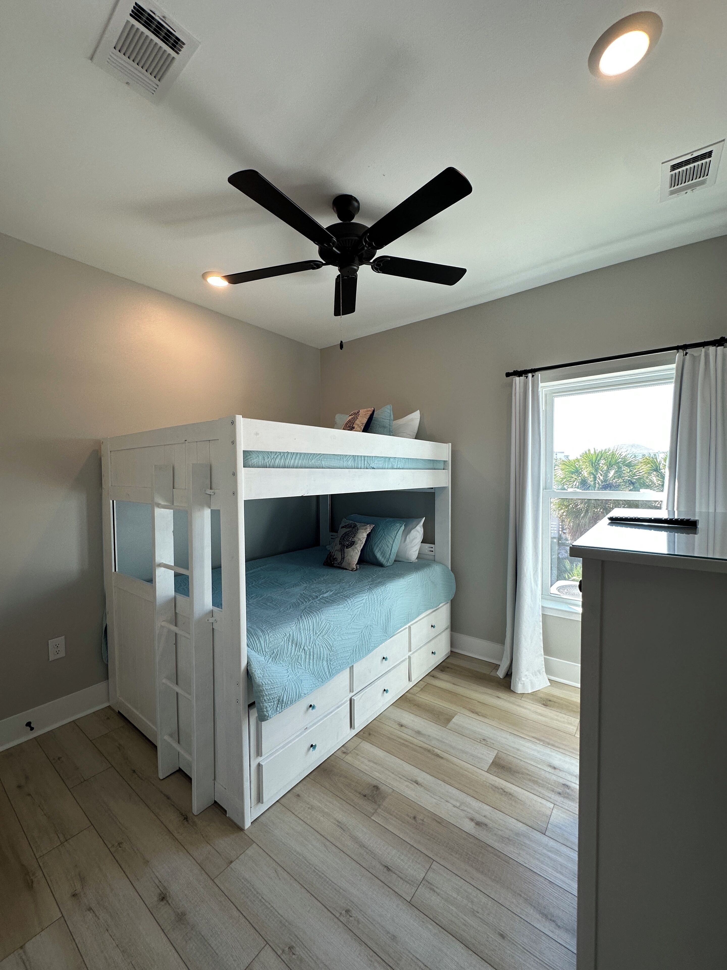 A bunk bed with a desk and a ceiling fan in a room with wooden flooring and a window with curtains.