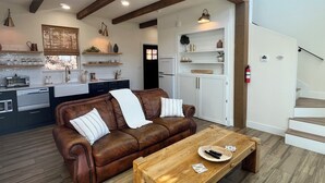 A modern living room with a brown leather sofa, wooden coffee table, and a kitchen area with white cabinets and a black refrigerator. - Travis Treehouse, Private Couples Getaway  (Austin)