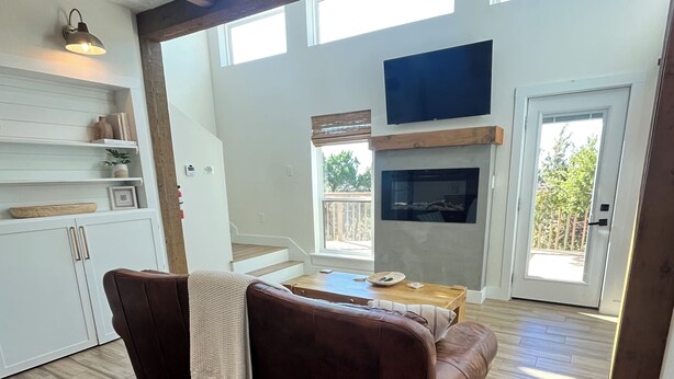 A spacious living area with a brown leather sofa, wooden flooring, a fireplace, and a large window providing natural light. - Travis Treehouse, Private Couples Getaway  (Austin)