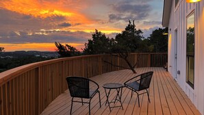A wooden deck with two chairs and a table, overlooking a sunset with trees and a distant landscape. - Travis Treehouse, Private Couples Getaway  (Austin)
