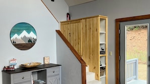 Uma sala de estar com um armário cinzento, uma escada de madeira, uma porta que dá para o exterior e uma obra de arte circular fixada na parede.