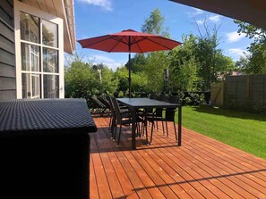 A wooden deck with a table and chairs, an umbrella, and a view of a green lawn and trees.
