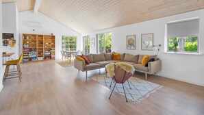 A spacious living room with a wooden ceiling, a sofa, a chair, a bookshelf, and a dining area.