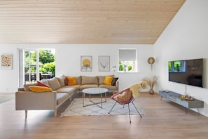 Ein modernes Wohnzimmer mit einem Ecksofa, einem runden Couchtisch, einem Fernseher und einem großen Fenster mit Blick auf Grünflächen.