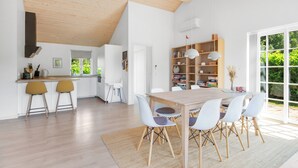 A modern kitchen with a wooden ceiling, a dining area with a wooden table and white chairs, and a bookshelf with various items.