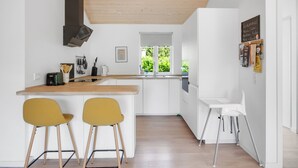 A modern kitchen with white cabinetry, a wooden ceiling, and a window with a view of greenery. There are two yellow chairs and a white chair.