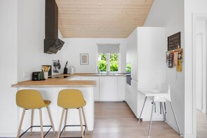 Eine moderne Küche mit weißen Schränken, einer Holzbalkendecke und einem Fenster mit Blick auf Grünflächen. Zwei gelbe Stühle und ein weißer Stuhl sind vorhanden.