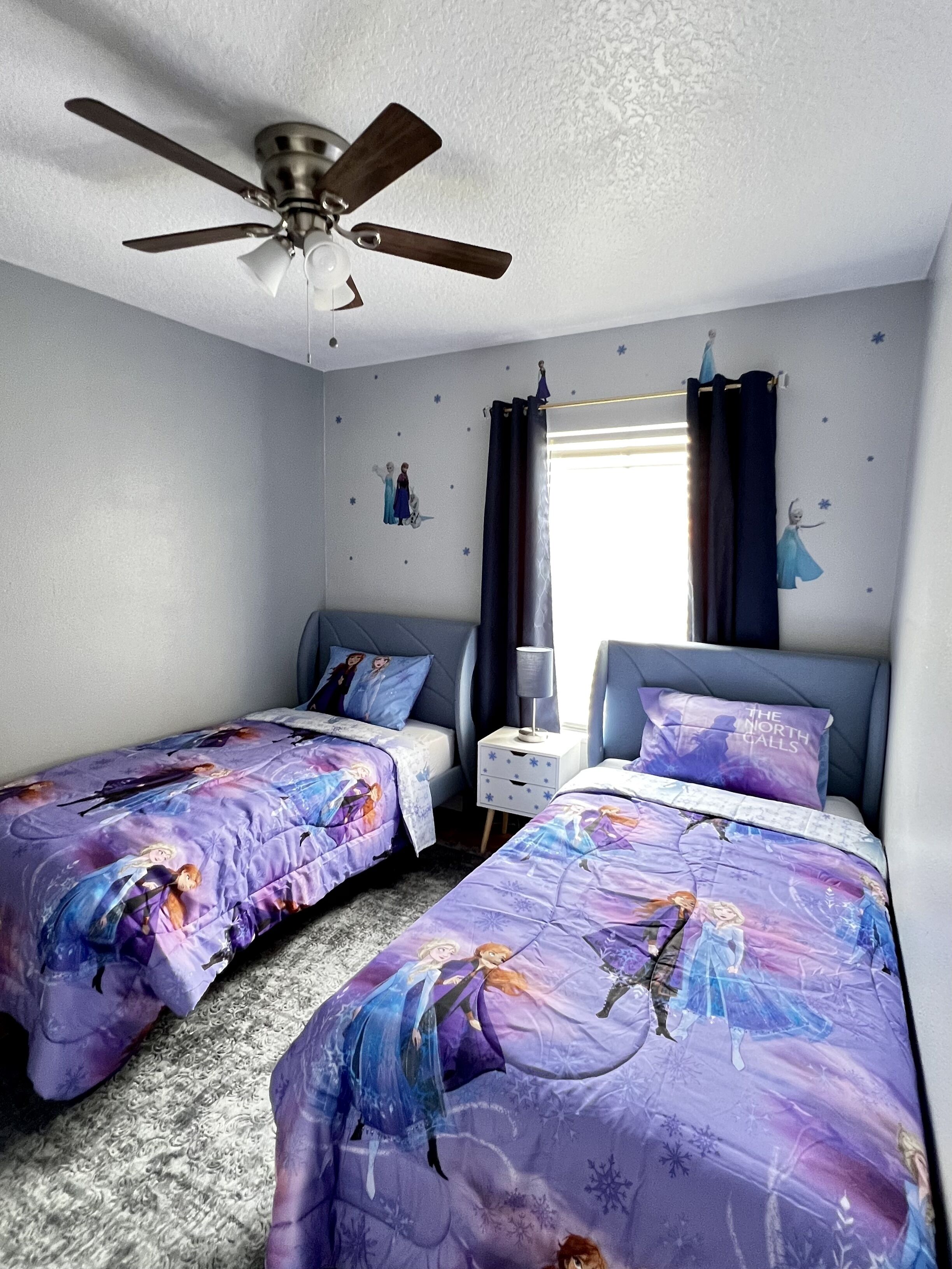 A bedroom with two beds, a ceiling fan, a nightstand, and a window with curtains.