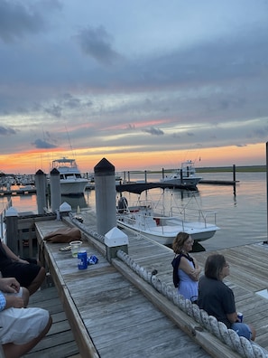 A wooden dock with people sitting, overlooking a marina with boats and a sunset in the background.