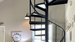 A modern interior with a spiral staircase, a wicker chair, a small table, and a wall-mounted lamp. - Blessed Rest (Edisto Island)