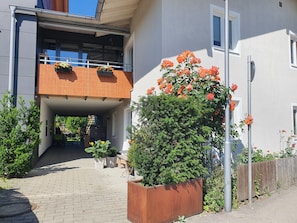 Une entrée d’immeuble avec un passage couvert, des plantes en pot et une clôture métallique.