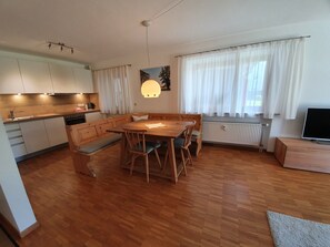 Une cuisine moderne avec des meubles en bois et un four encastré. On y trouve un coin repas avec une table et des bancs en bois. La pièce est dotée de grandes fenêtres avec des rideaux blancs.