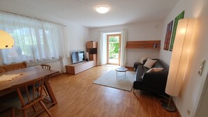 A modern living room with a wooden dining table, a black sofa, a TV on a wooden stand, and a large window with sheer curtains. - Ferienwohnung 54qm mit Sep. Schlafzimmer und Terrasse (Prien am Chiemsee)