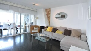 A modern living room with a glass coffee table, a sofa with cushions, a wicker chair, and a large mirror on the wall. - Rental in La Almadrava beach, Roses. Apartment with sea views, private terrace, equipped kitchen. Book now! (Roses)