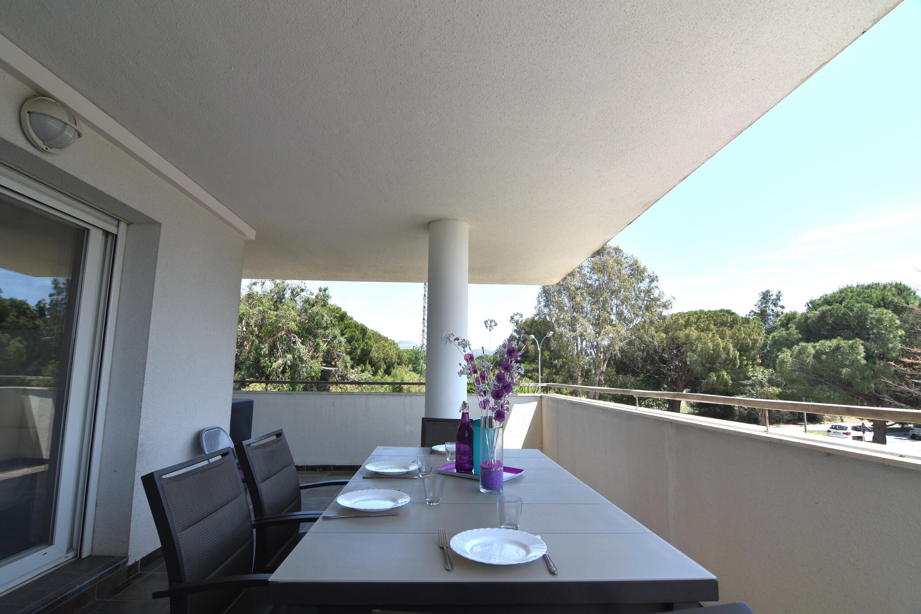 A balcony with a table set for two, featuring a vase with flowers, a bottle, and a glass.